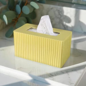 May include: A yellow tissue box with vertical ribbed detailing. The box is rectangular with a rectangular opening at the top, and white tissues are visible. The box is on a white marble surface.