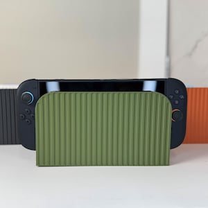 May include: A black handheld gaming console is displayed in front of three ribbed, arched stands. The stands are in shades of gray, olive green, and orange. The console has black buttons and a black screen.