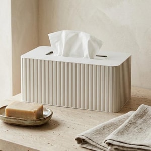 Peut inclure: Scène de salle de bain aux tons neutres avec une boîte à mouchoirs, un porte-savon et une serviette pliée. La boîte à mouchoirs est rectangulaire avec des détails verticaux côtelés. Un pain de savon repose sur un petit porte-savon ovale.