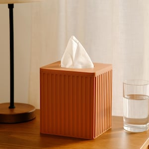 May include: A square, orange tissue box with vertical ridged detailing. White tissues are visible at the top. The box sits on a wooden surface next to a glass of water and a lamp.