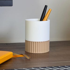 May include: A white ceramic pencil holder with a wood-grain base. The holder has two pens inside, one black and one yellow.