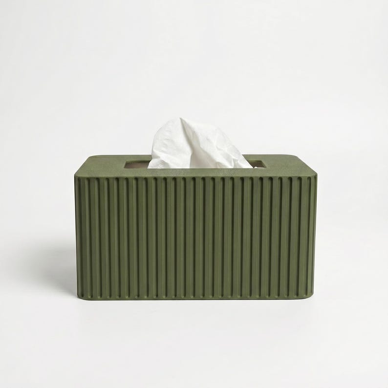 May include: A rectangular olive green tissue box cover featuring a vertical ribbed texture on all visible sides. The top surface has a central rectangular opening from which a single white paper tissue is protruding upwards in a folded shape.
