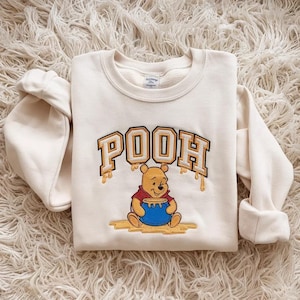 Peut inclure: Un sweat-shirt à col rond de couleur crème avec une broderie représentant le personnage Winnie l'ourson assis tenant un pot de miel bleu. Le mot POOH est brodé dans une police universitaire jaune et blanche avec des détails de miel coulant.