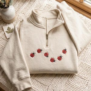 May include: A cream-colored quarter-zip sweatshirt featuring an embroidered pattern of small red ladybugs with black spots across the chest. The garment has a high collar, long sleeves with ribbed cuffs, and a metallic zipper closure at the neckline.