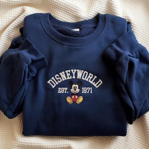 Puede incluir: Sudadera azul marino con el texto "DISNEYWORLD" en letras blancas arqueadas, con "EST. 1971" debajo. Un gráfico de Mickey Mouse está centrado debajo del texto. La sudadera está doblada sobre una superficie texturizada de color crema.