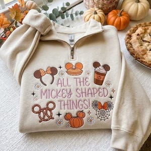Pode incluir: Uma camisola bege com fecho de correr curto, apresentando ícones bordados incluindo orelhas de rato, uma abóbora, um pretzel e um copo de café, com o texto "ALL THE MICKEY SHAPED THINGS!" rodeado por pequenos detalhes de flores e estrelas.