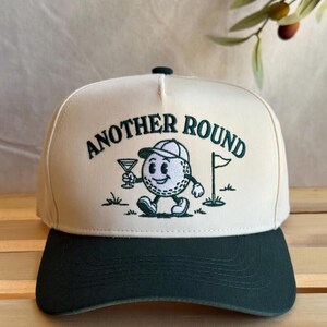 Op de afbeelding: Een crèmekleurige baseballcap met een donkergroene klep. De cap heeft de tekst "ANOTHER ROUND" in het groen, samen met een cartoon golfbal karakter met een cocktail. Een kleine golfvlag is ook inbegrepen.