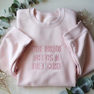 May include: A light pink sweatshirt with the embroidered text "THE BRIDE NEEDS A DIET COKE". The crew neck sweatshirt has long sleeves and is made of a soft material. Perfect for a bachelorette party or bridal shower.