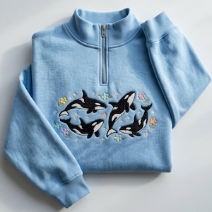 May include: A light blue quarter-zip sweatshirt featuring an embroidered design of four black and white orcas swimming among small, colourful pastel flowers and tiny four-pointed stars. The garment has a ribbed collar and long sleeves with ribbed cuffs.