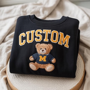May include: A black crewneck sweatshirt featuring the word CUSTOM embroidered in yellow block letters. Below the text is an embroidered brown teddy bear wearing a dark blue sweater with a yellow letter M on the front and light tan paw pads.