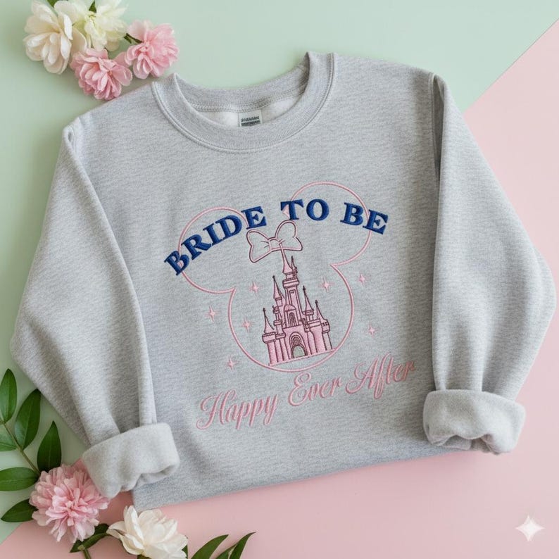 Puede incluir: Una sudadera gris jaspeado de cuello redondo con un dise&ntilde;o bordado de la silueta de un castillo dentro del contorno de una cabeza de Mickey Mouse. El texto dice BRIDE TO BE en azul oscuro y Happy Ever After en rosa, con un lazo peque&ntilde;o.