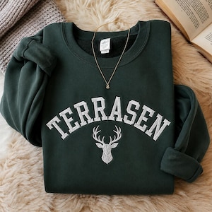 May include: A dark forest green crewneck sweatshirt featuring the word TERRASEN embroidered in white block letters in an arched formation above a detailed white embroidered stag head graphic centred on the chest area of the garment.