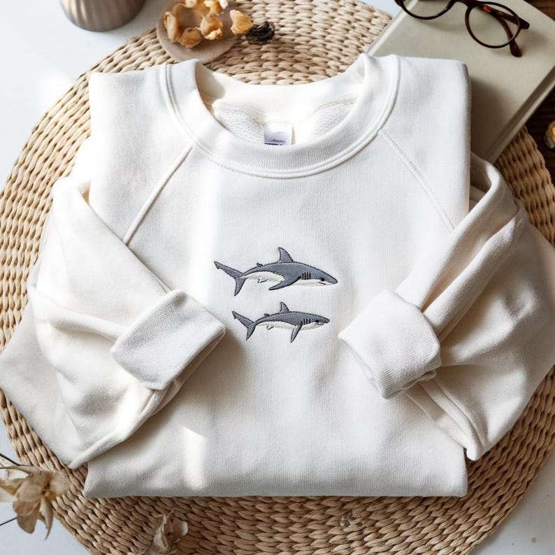 K&ouml;nnte beinhalten: Ein cremefarbenes Sweatshirt mit Rundhalsausschnitt und einer Stickerei von zwei grauen Haien, die &uuml;bereinander in der Mitte der Brust platziert sind. Das Kleidungsst&uuml;ck hat lange &Auml;rmel mit gerippten B&uuml;ndchen und einen gerippten Halsausschnitt.