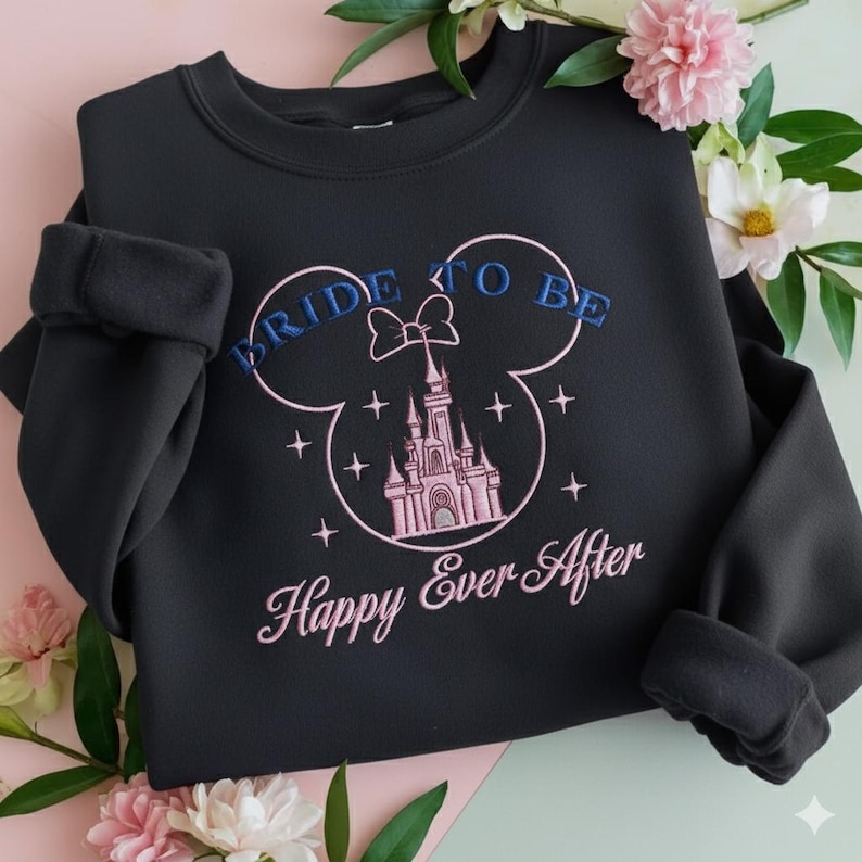 Puede incluir: Una sudadera negra de cuello redondo con un dise&ntilde;o bordado de un castillo dentro del contorno de unas orejas de Mickey Mouse. El texto dice BRIDE TO BE en azul y Happy Ever After en rosa, decorado con peque&ntilde;as estrellas y un lazo sobre el castillo.