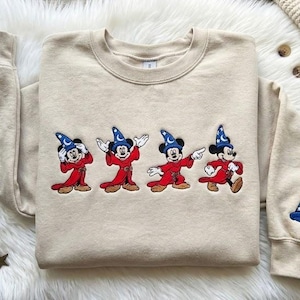 May include: Beige sweatshirt featuring an embroidered design of Mickey Mouse in a sorcerer's outfit. The design is repeated across the front, with a single sorcerer's hat on the left sleeve. The sweatshirt is laid on a white fluffy surface.