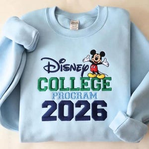 May include: A light blue crewneck sweatshirt featuring embroidered text that reads Disney COLLEGE PROGRAM 2026. The design includes a colorful embroidered graphic of Mickey Mouse positioned above the green text. The sweatshirt has long sleeves.