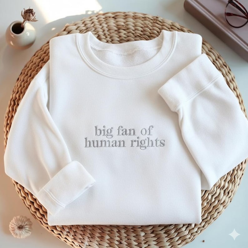 May include: A white sweatshirt with the phrase "big fan of human rights" printed in gray. The long-sleeved top is folded on a woven wicker mat. The cuffs are folded up. A small white vase and a book are in the background.