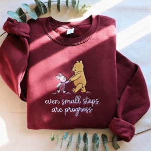 May include: Burgundy sweatshirt featuring an embroidered image of Winnie the Pooh and Piglet holding hands. The text "even small steps are progress" is below the characters. The sleeves are cuffed.