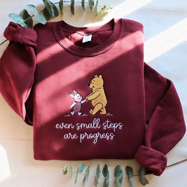 May include: Burgundy sweatshirt featuring an embroidered image of Winnie the Pooh and Piglet holding hands. The text "even small steps are progress" is below the characters. The sleeves are cuffed.