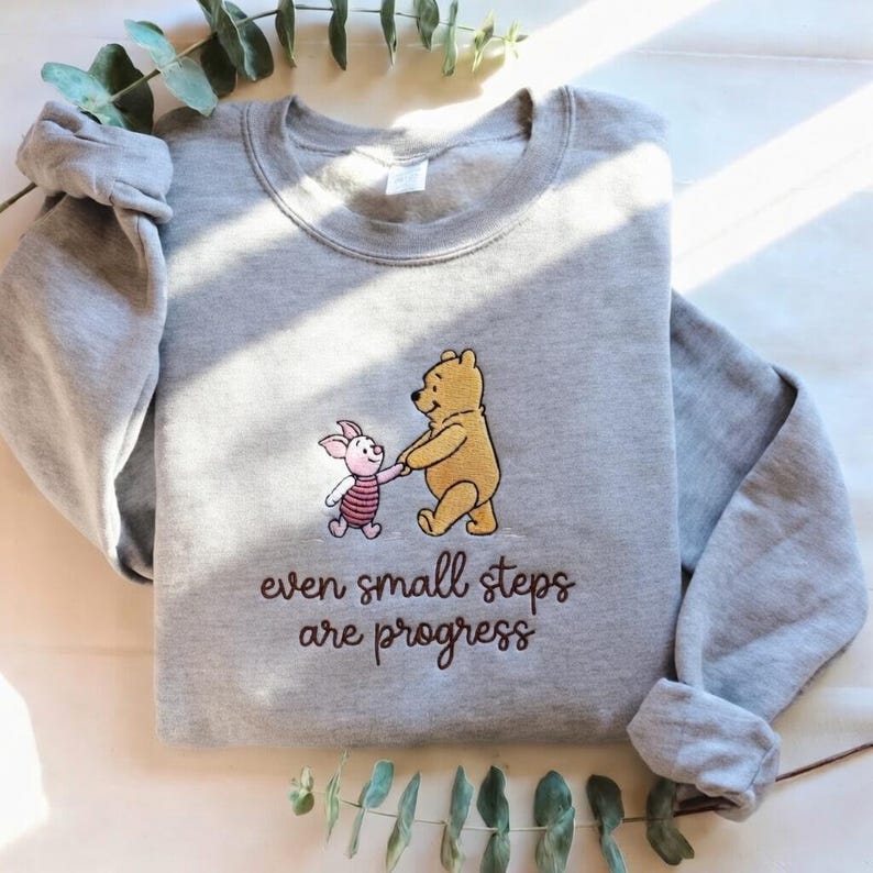 May include: A gray sweatshirt featuring an embroidered design of Winnie the Pooh and Piglet holding hands. The text "even small steps are progress" is below the characters. The sweatshirt has long sleeves.