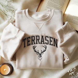 May include: A cream-coloured sweatshirt featuring the word "TERRASEN" in black arched lettering and a black deer head graphic. The sweatshirt has a crew neck and long sleeves. A lit candle and an open book are in the background.