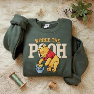 May include: A green Winnie the Pooh sweatshirt with the words "WINNIE THE POOH" in yellow and white block letters. The image features a cartoon of Winnie the Pooh reaching into a honey pot. The word "HUNNY" is on the honey pot.