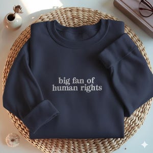 May include: Navy blue sweatshirt with the embroidered text "big fan of human rights." The sweatshirt is folded on a woven, circular mat. A small white vase with a dried flower and a decorative item are in the background.