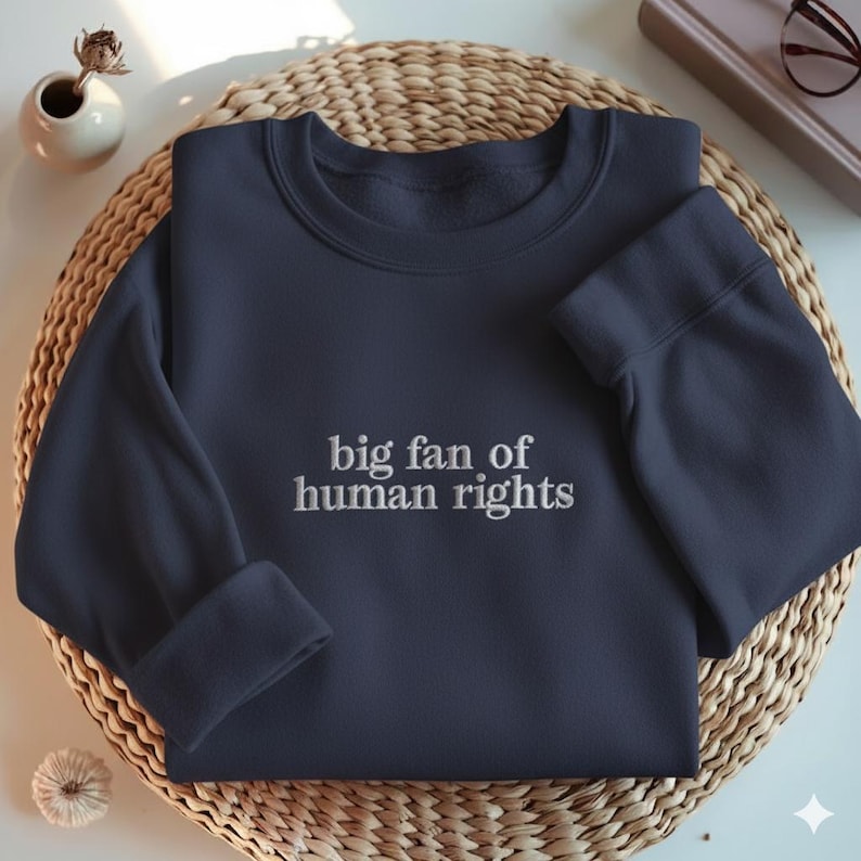 May include: Navy blue sweatshirt with the embroidered text "big fan of human rights." The sweatshirt is folded on a woven, circular mat. A small white vase with a dried flower and a decorative item are in the background.