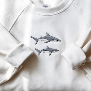K&ouml;nnte beinhalten: Ein cremefarbenes Sweatshirt mit Rundhalsausschnitt und einer Stickerei von zwei grauen Haien, die &uuml;bereinander in der Mitte der Brust platziert sind. Das Kleidungsst&uuml;ck hat lange &Auml;rmel mit gerippten B&uuml;ndchen und einen gerippten Halsausschnitt mit Etikett.