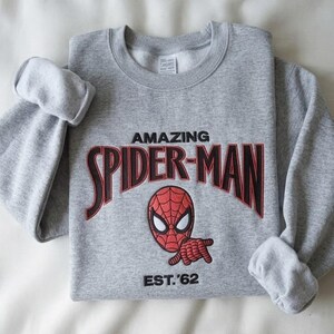 May include: A heather gray crewneck sweatshirt featuring embroidered text that reads AMAZING SPIDER-MAN in black with red outlines. Below the text is an embroidered red and black Spider-Man mask and the black embroidered text EST. '62.