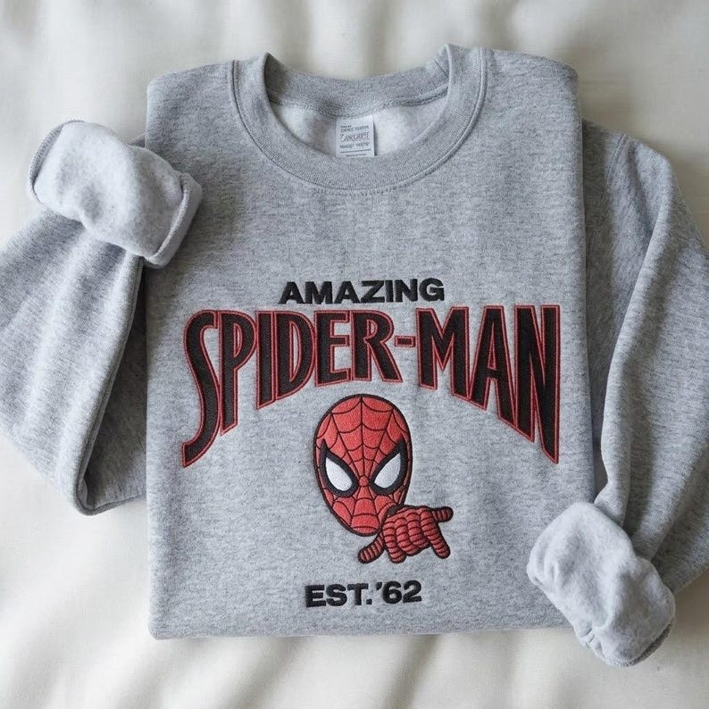 May include: A heather gray crewneck sweatshirt featuring embroidered text that reads AMAZING SPIDER-MAN in black with red outlines. Below the text is an embroidered red and black Spider-Man mask and the black embroidered text EST. '62.
