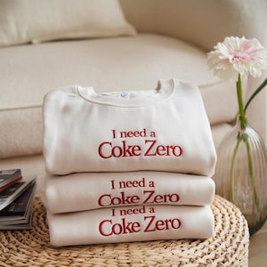 May include: A stack of three cream-coloured sweatshirts with the text "I need a Coke Zero" embroidered in red on each.