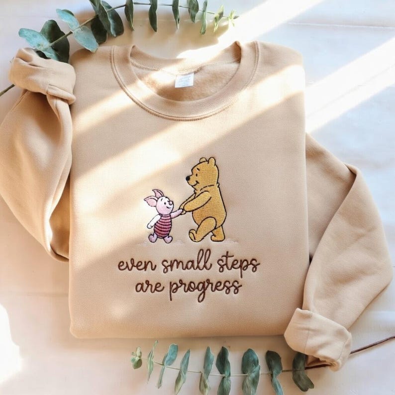 May include: A tan sweatshirt featuring an embroidered image of Winnie the Pooh and Piglet holding hands. The text "even small steps are progress" is embroidered below the characters. The sleeves are rolled up.