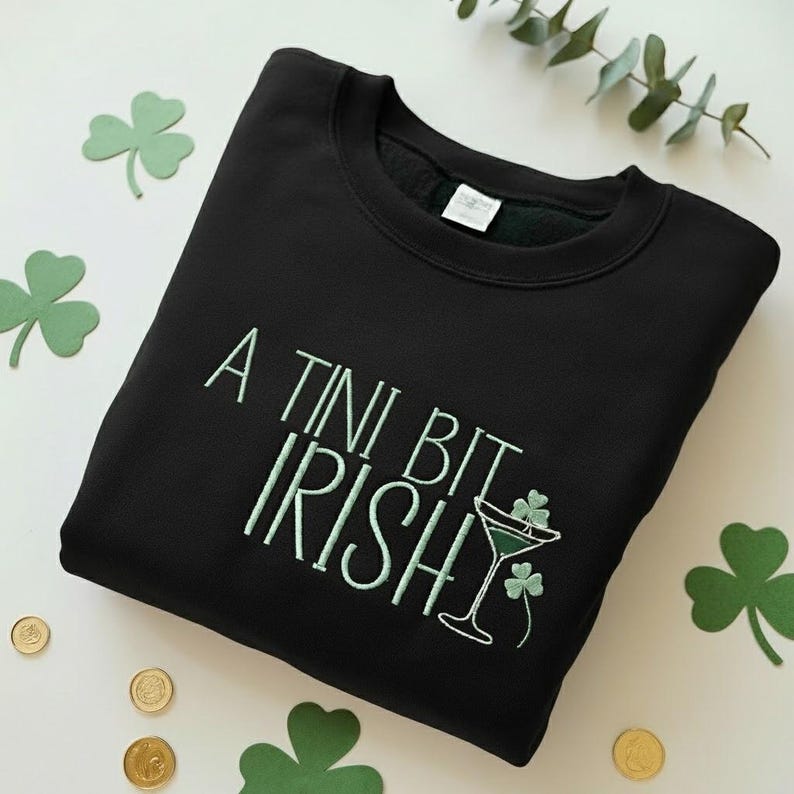 May include: Black sweatshirt with the embroidered text "A Tini Bit Irish" in light green. The design includes a martini glass with shamrocks. The image also features shamrock cutouts and gold coins, suggesting a St. Patrick's Day theme.