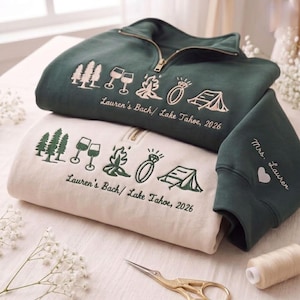 May include: Two quarter-zip sweatshirts, one dark green and one cream, featuring embroidered icons of pine trees, wine glasses, a campfire, a ring, and a tent. Text reads "Lauren's Bach/ Lake Tahoe, 2026". The green sleeve shows "Mrs. Lauren" with a heart.