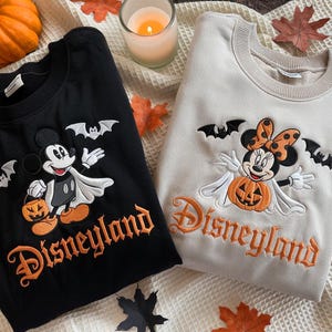 May include: A black sweatshirt and a beige sweatshirt, both featuring embroidered designs of Mickey and Minnie Mouse dressed as ghosts with pumpkins and bats. The word Disneyland is embroidered in orange gothic-style lettering on each garment.