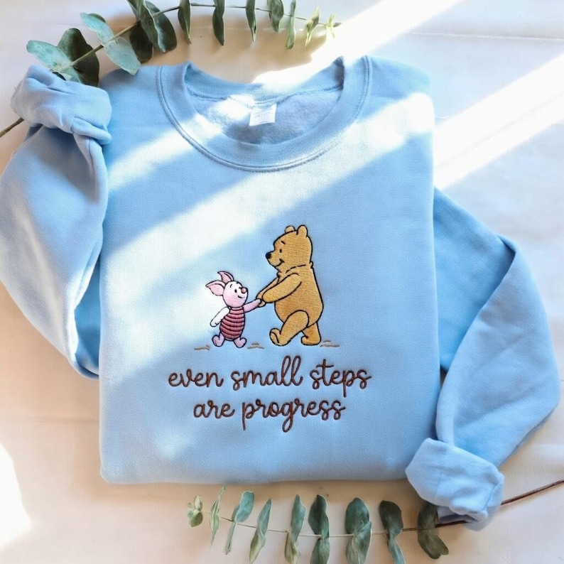 May include: Light blue sweatshirt featuring an embroidered design of Winnie the Pooh and Piglet holding hands. The text "even small steps are progress" is below the characters. The long-sleeved top is folded.