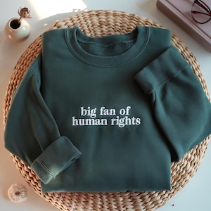 May include: A dark green sweatshirt with the phrase "big fan of human rights" embroidered in white. The sweatshirt is folded on a woven wicker mat. A small white vase with a dried flower and a book with glasses are in the background.