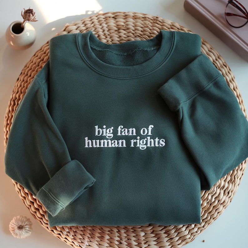 May include: A dark green sweatshirt with the phrase "big fan of human rights" embroidered in white. The sweatshirt is folded on a woven wicker mat. A small white vase with a dried flower and a book with glasses are in the background.