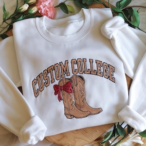 Peut inclure: Sweat-shirt blanc avec le texte "CUSTOM COLLEGE" en broderie marron. Le sweat-shirt présente une paire de bottes de cow-boy marron avec un nœud rouge.