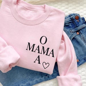 May include: A light pink sweatshirt with the text "O MAMA A ♡" in black. The sweatshirt is folded over a pair of blue denim jeans. The sweatshirt has long sleeves and a crew neck.