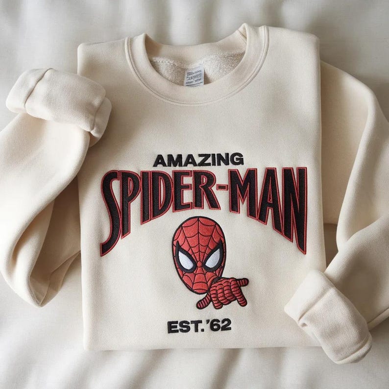 May include: A cream-colored crewneck sweatshirt featuring an embroidered graphic of the Spider-Man mask and hand. The text reads AMAZING SPIDER-MAN in black with red outlines, and EST. '62 is embroidered in black below the character graphic.