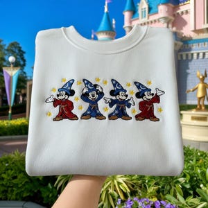 May include: White sweatshirt with an embroidered design of Mickey Mouse in wizard outfits. The design shows Mickey in various poses, wearing blue and red robes and wizard hats, with gold stars. A fun and whimsical design.