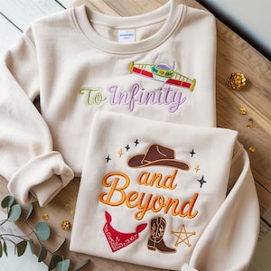 Puede incluir: Dos sudaderas beige con diseños bordados. Una presenta una nave espacial y el texto "To Infinity" en verde y morado. La otra tiene un sombrero de vaquero, botas y el texto "and Beyond" en naranja.
