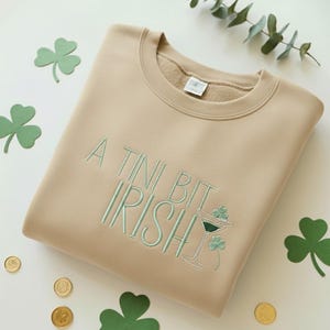 May include: A tan sweatshirt with the embroidered phrase "A Tini Bit Irish" in light green. The design includes a martini glass with shamrocks. Green shamrock cutouts and gold coins are scattered around the sweatshirt.