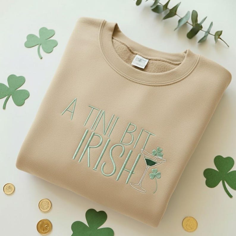 May include: A tan sweatshirt with the embroidered phrase "A Tini Bit Irish" in light green. The design includes a martini glass with shamrocks. Green shamrock cutouts and gold coins are scattered around the sweatshirt.