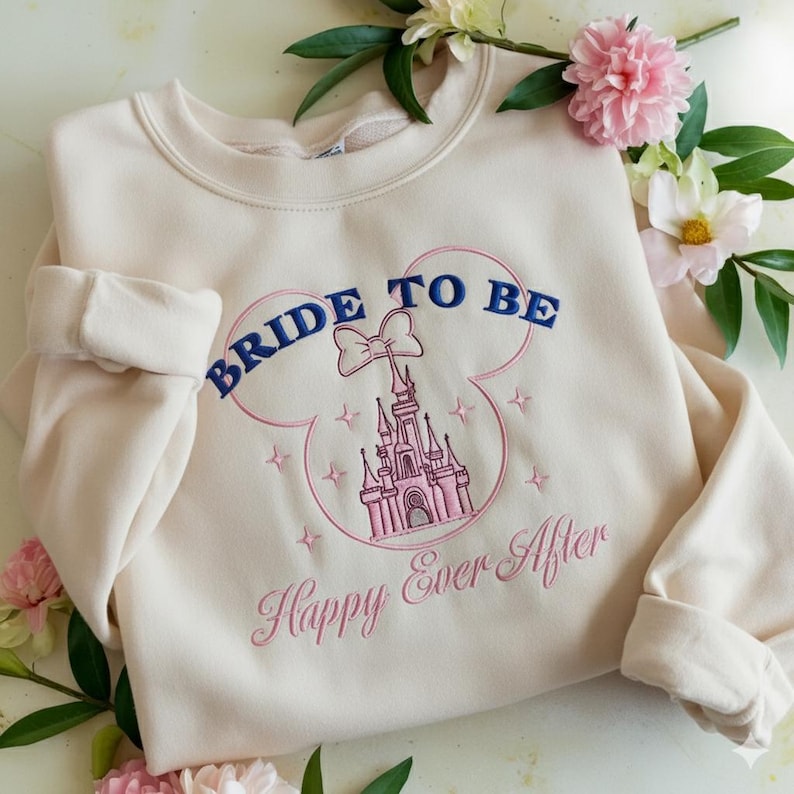 Puede incluir: Una sudadera de cuello redondo color crema con un dise&ntilde;o bordado de la silueta de un castillo rosa dentro del contorno de unas orejas de Mickey Mouse. El texto dice BRIDE TO BE en azul oscuro y Happy Ever After en cursiva rosa.
