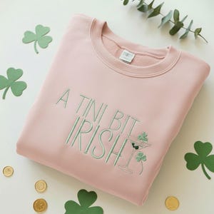 May include: A light pink sweatshirt with the embroidered phrase "A Tini Bit Irish" in green. The design includes a martini glass with shamrocks. Green shamrock cutouts and gold coins are scattered around the sweatshirt.