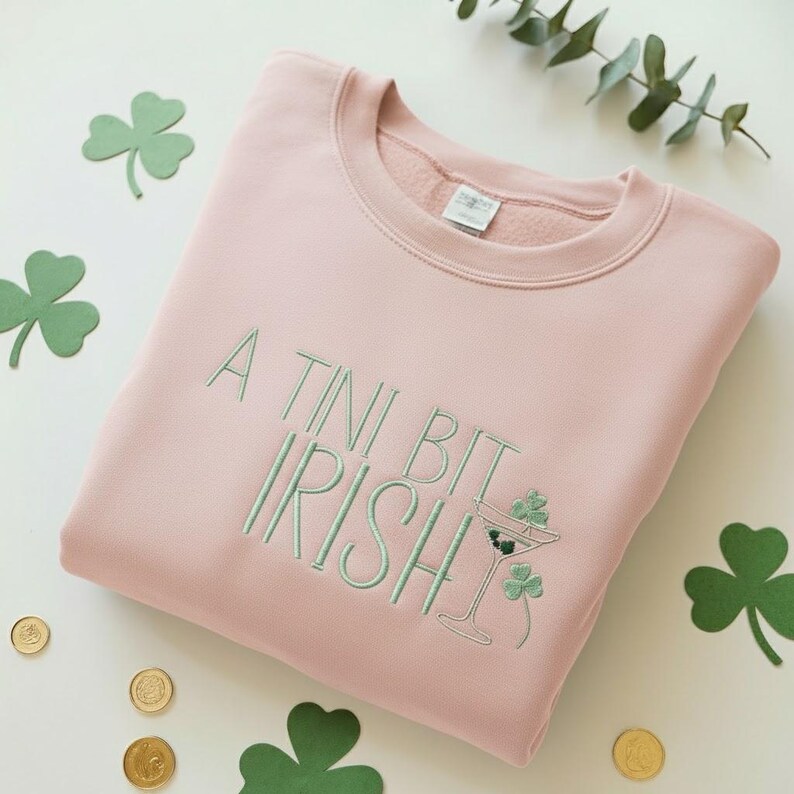 May include: A light pink sweatshirt with the embroidered phrase "A Tini Bit Irish" in green. The design includes a martini glass with shamrocks. Green shamrock cutouts and gold coins are scattered around the sweatshirt.