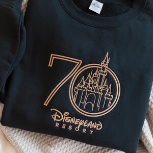 Puede incluir: Sudadera negra con dise&ntilde;o bordado en dorado. El dise&ntilde;o presenta el n&uacute;mero 70, un castillo dentro de un c&iacute;rculo y las palabras "Disneyland Resort". La sudadera est&aacute; doblada sobre una manta de color neutro.